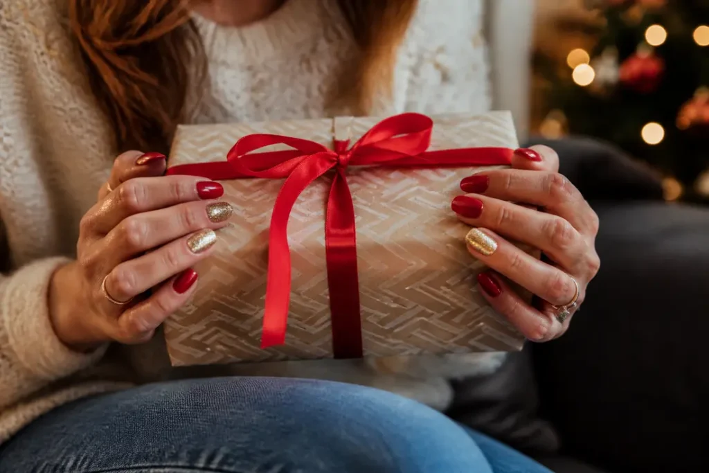 Mains élégantes avec une manucure noël rouge et doré tenant un cadeau de fêtes.