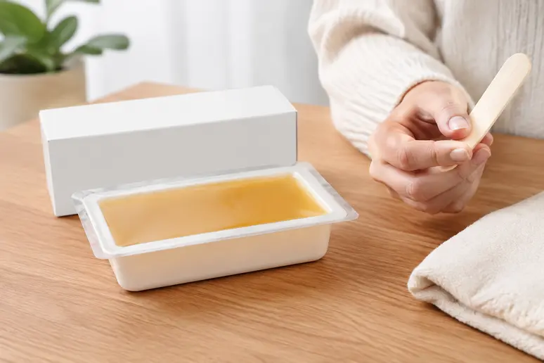Boîte de cire dépilatoire posée sur une surface en bois avec une spatule en bois